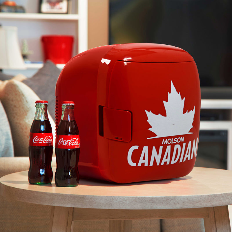 Lifestyle image of Molson Canadian 12 can cooler/warmer, closed, on light beige side table with two glass bottles of pop beside it and a couch, bookcase, and TV in the background